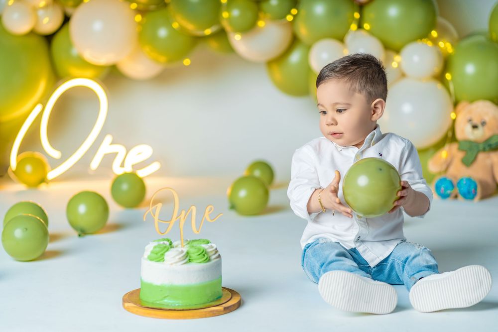 Curious baby boy examines green balloon beside first birthday cake with green and gold balloon arch.