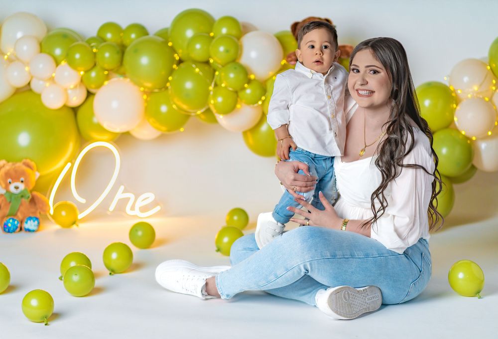 A mother sits on a white floor holding her toddler, surrounded by green and white balloons with a neon One sign.