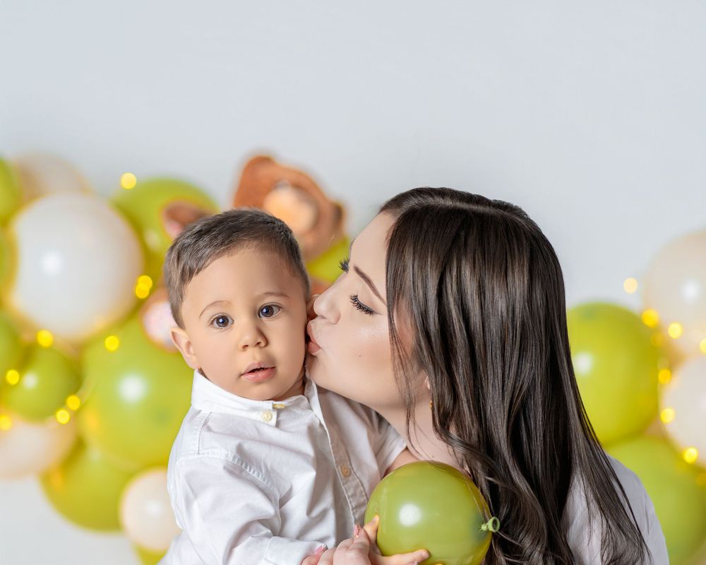 A mother tenderly kisses her baby boy cheek at a festive green and gold balloon backdrop celebration.