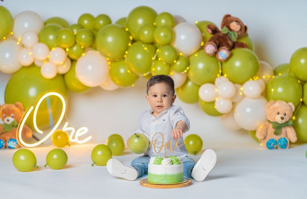 Baby's first birthday cake smash with green balloons, teddy bears, and "One" sign backdrop.