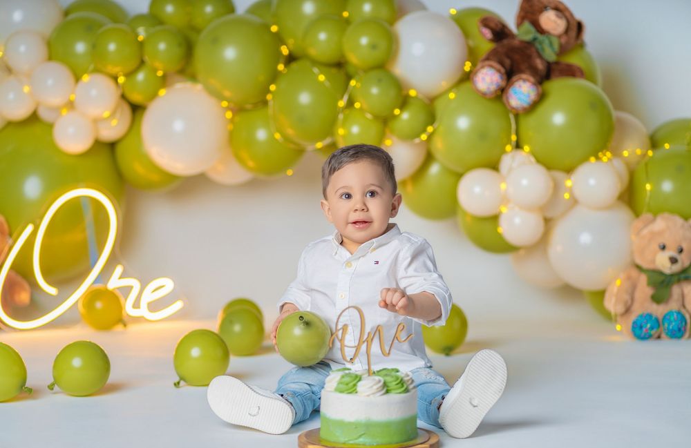 Baby boy sits before a cake smash setup with green and white balloons and teddy bears for a first birthday photo.