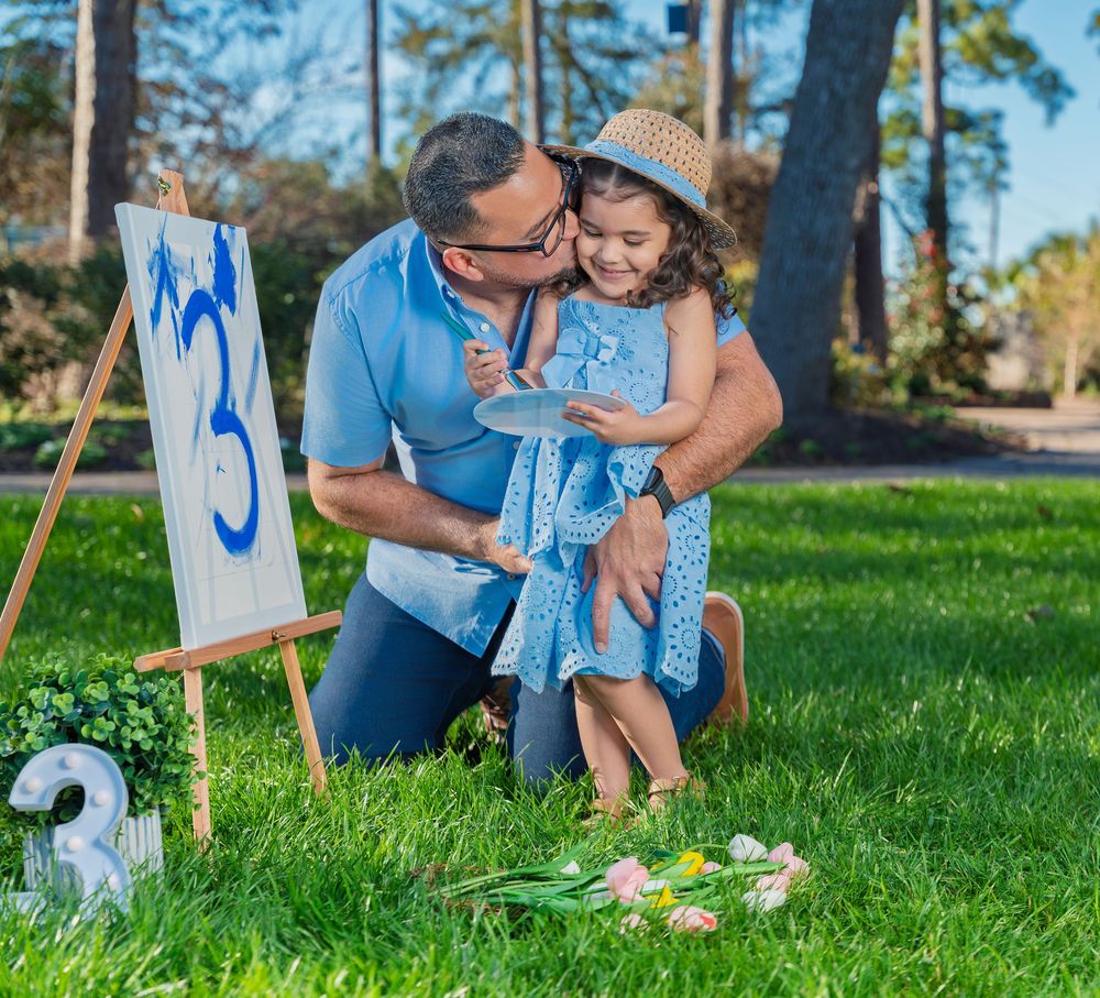 Father kneeling on grass kissing young girl in blue dress and straw hat near a number 3 birthday easel.