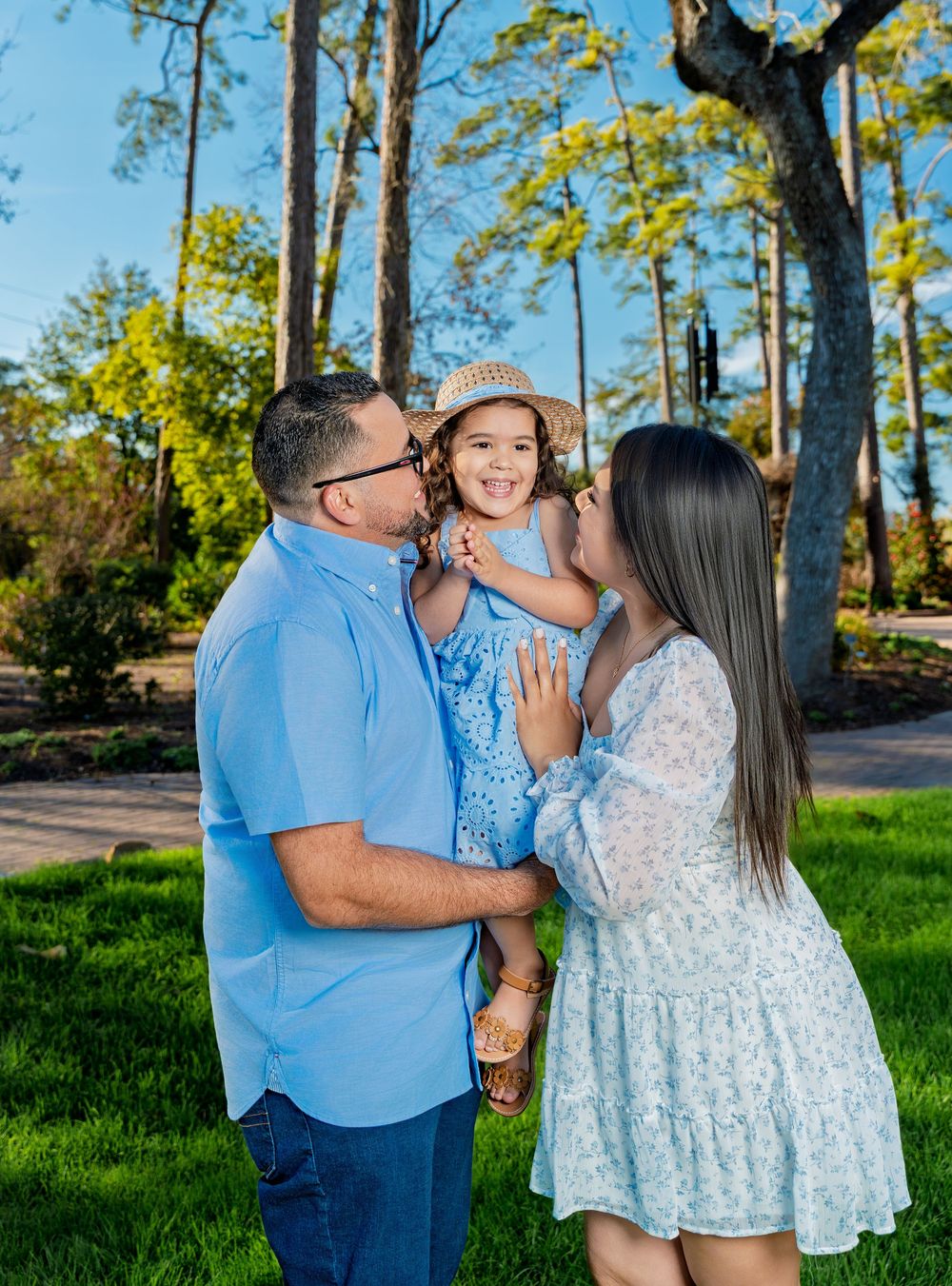 Parents kissing their smiling toddler daughter in blue dress and hat during outdoor birthday photo session.