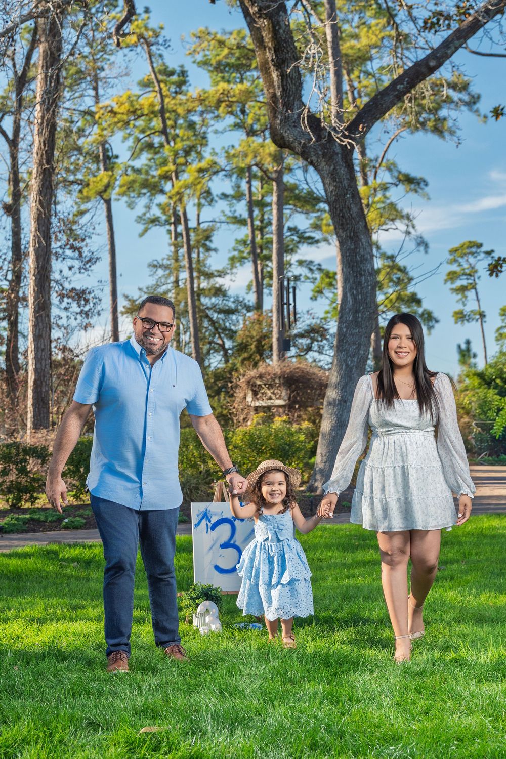 Family of three walking on green grass, parents holding toddler girl's hands, trees in sunny park background.
