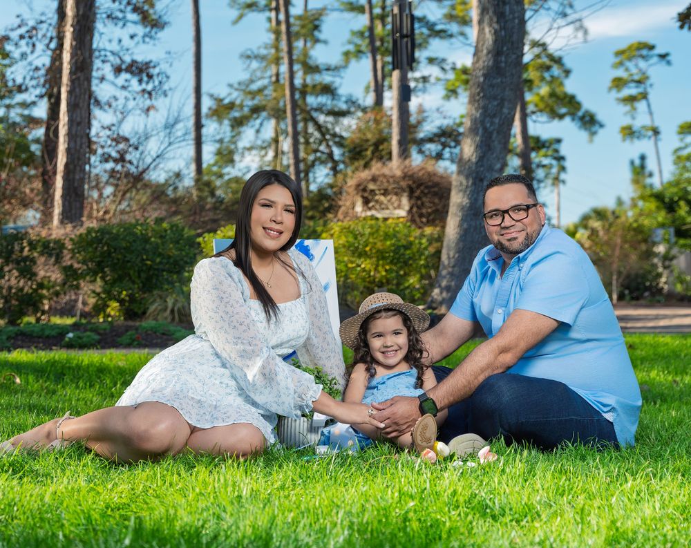 Happy family of three sitting on green grass outdoors with toddler girl in hat for birthday photos.