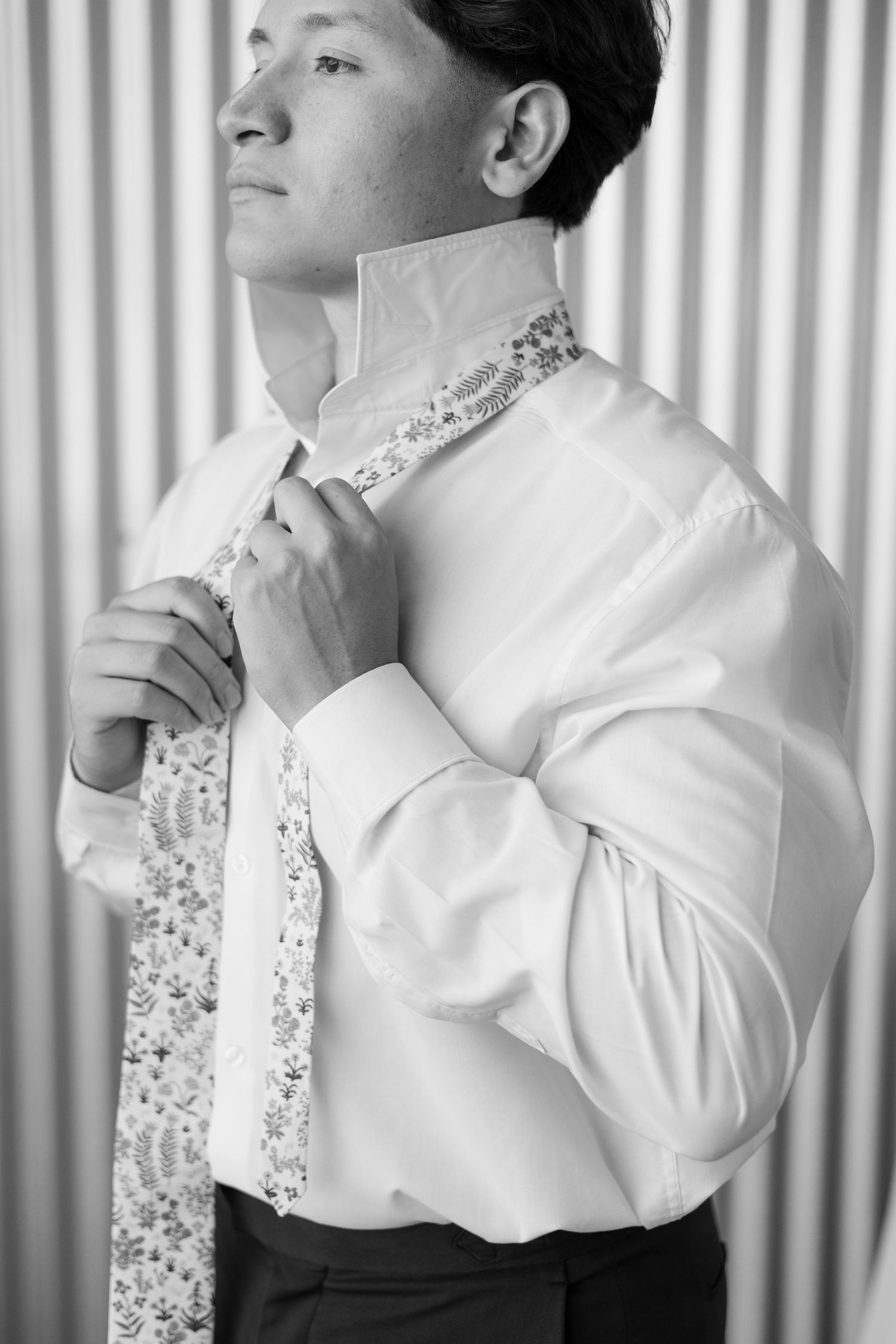 Black and white portrait of groom adjusting an embellished tie against striped background.