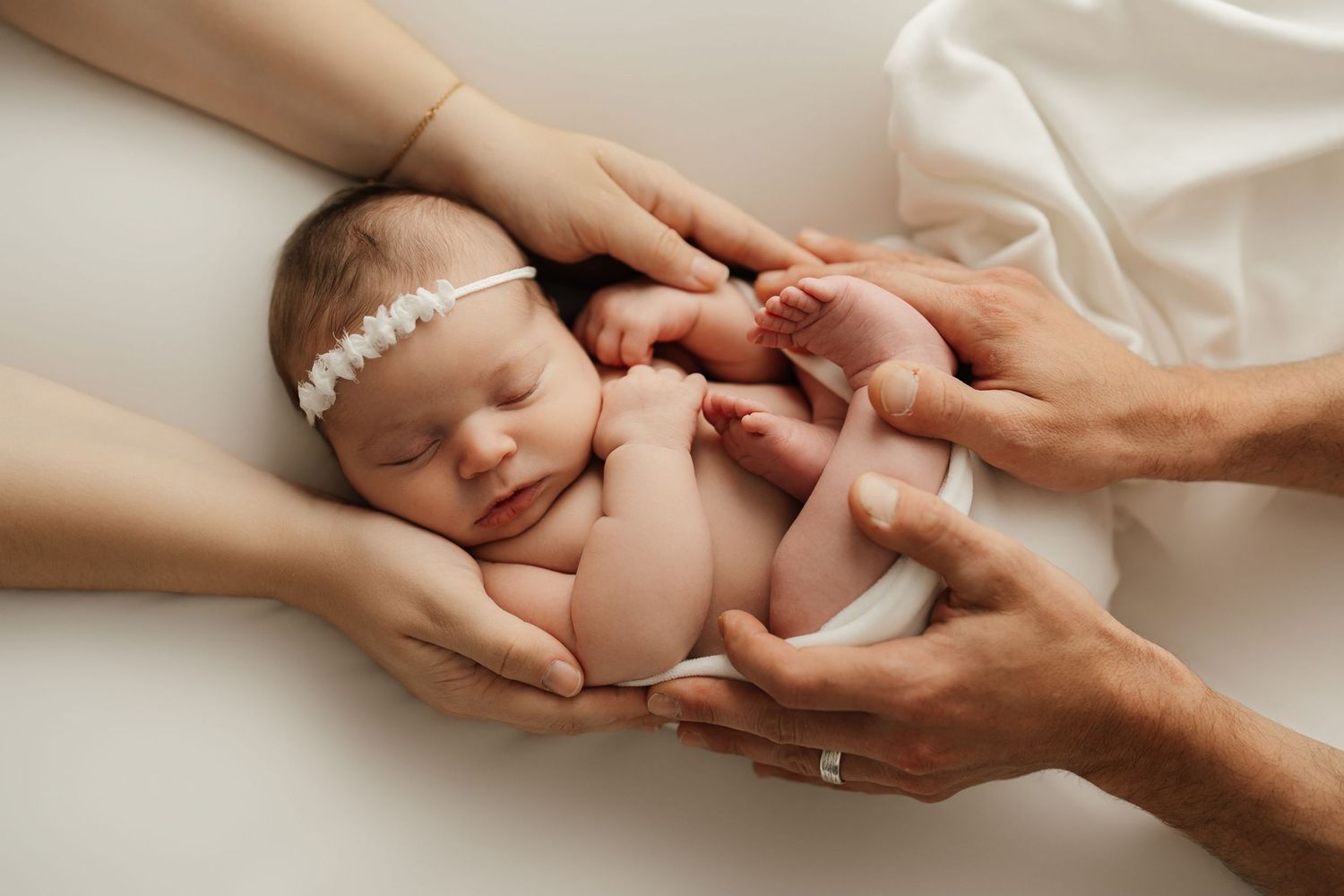 Gentle hands cradle a sleeping newborn baby wearing a delicate headband.