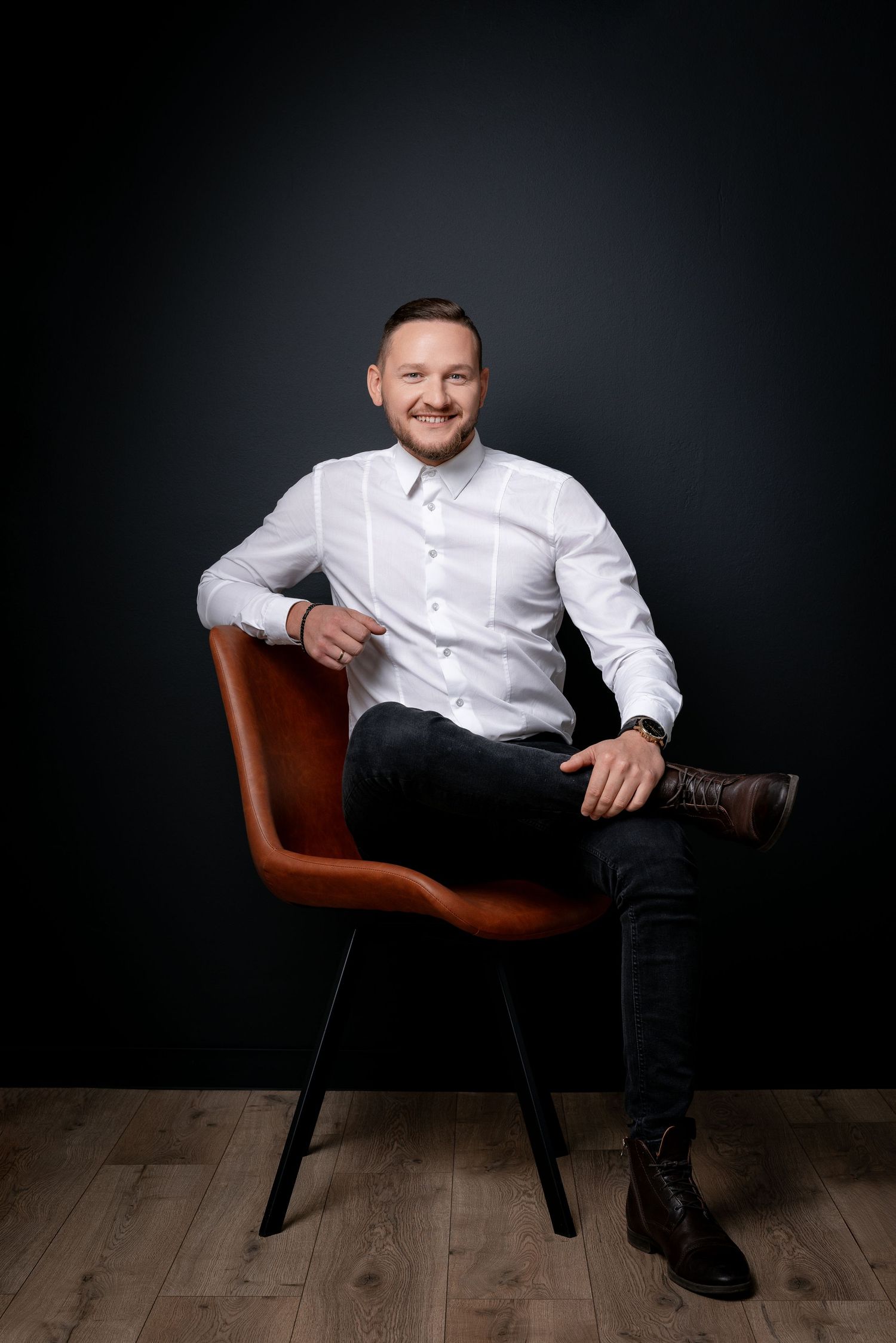 Professional portrait of someone in white shirt sitting on brown leather chair against dark background.