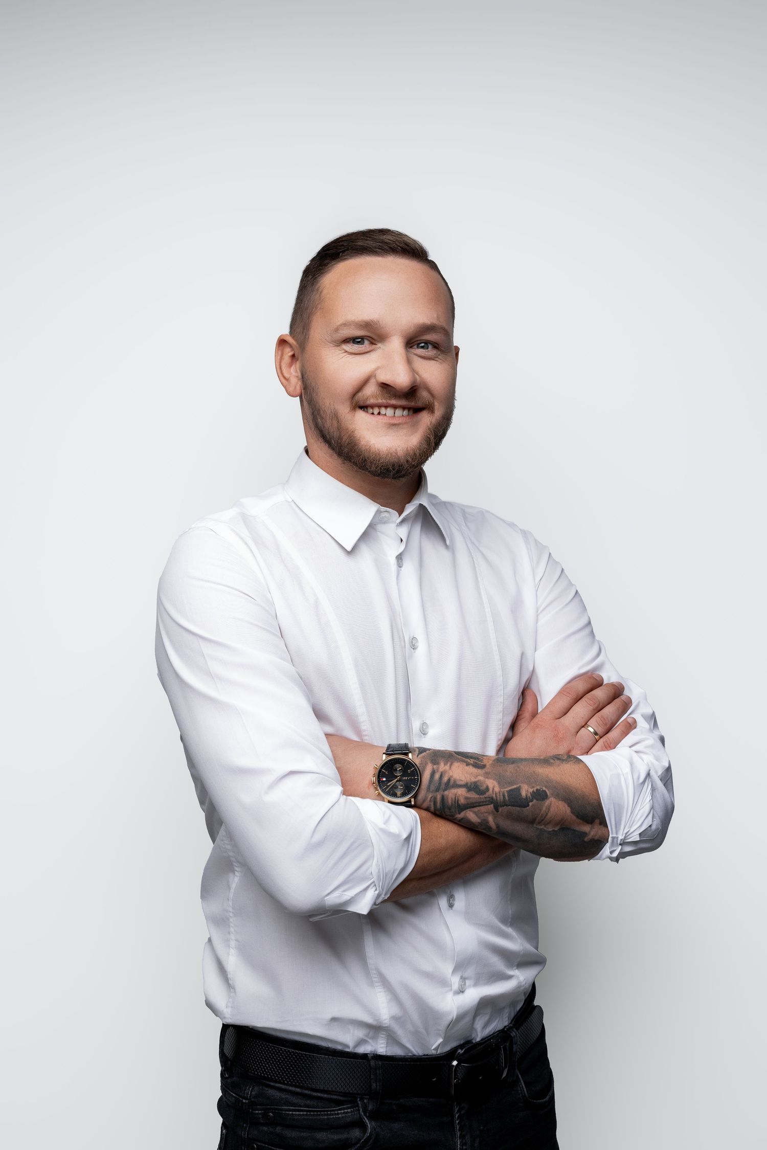 Professional portrait of person in white button-up shirt with tattoos and crossed arms against light background.