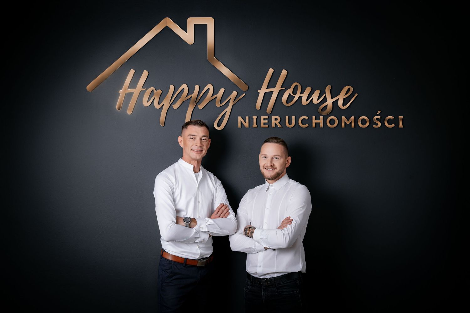 Two professionals pose in front of a dark wall with copper Happy House real estate logo and branding.