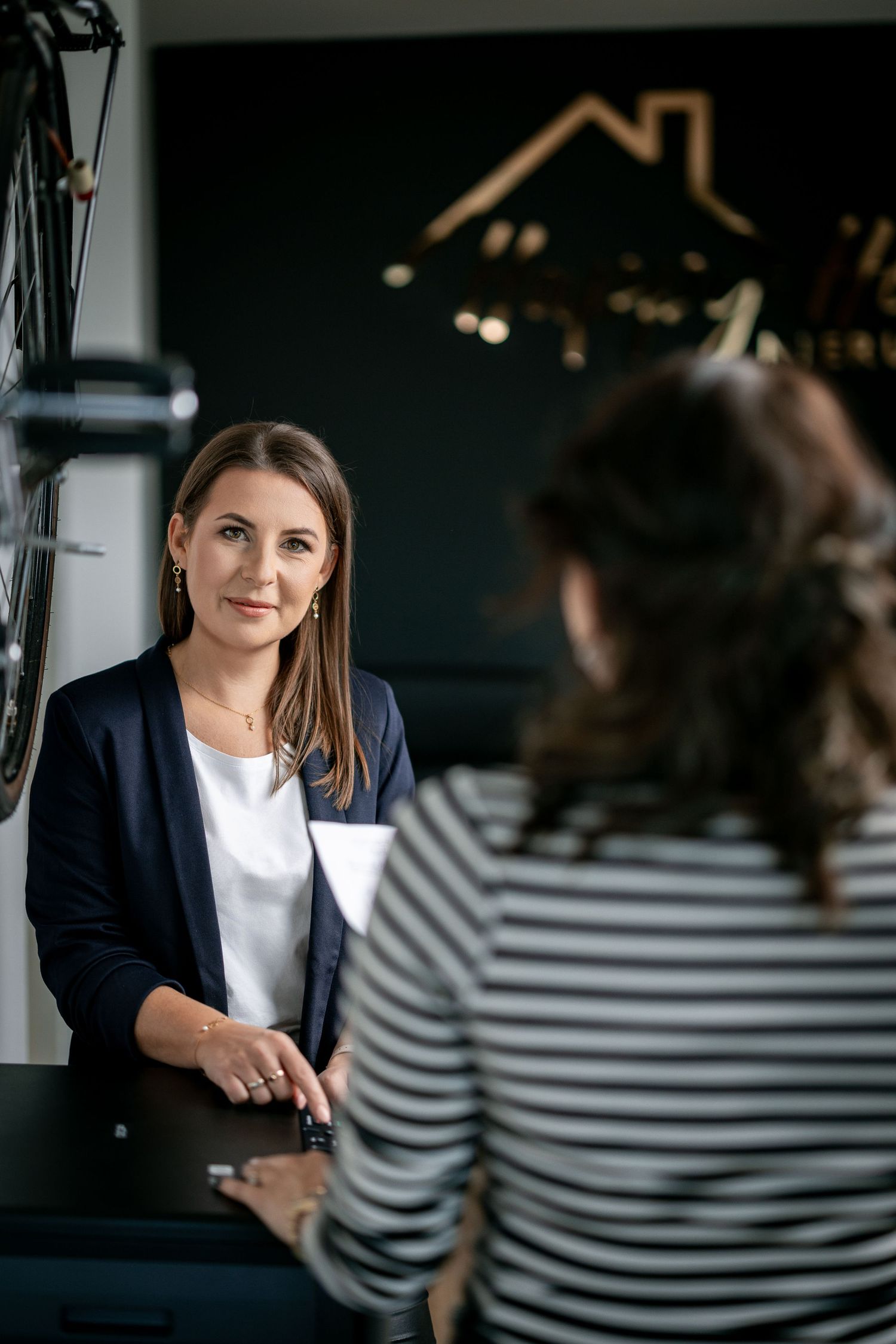 Professional business meeting between two people with house logo on dark wall in background.