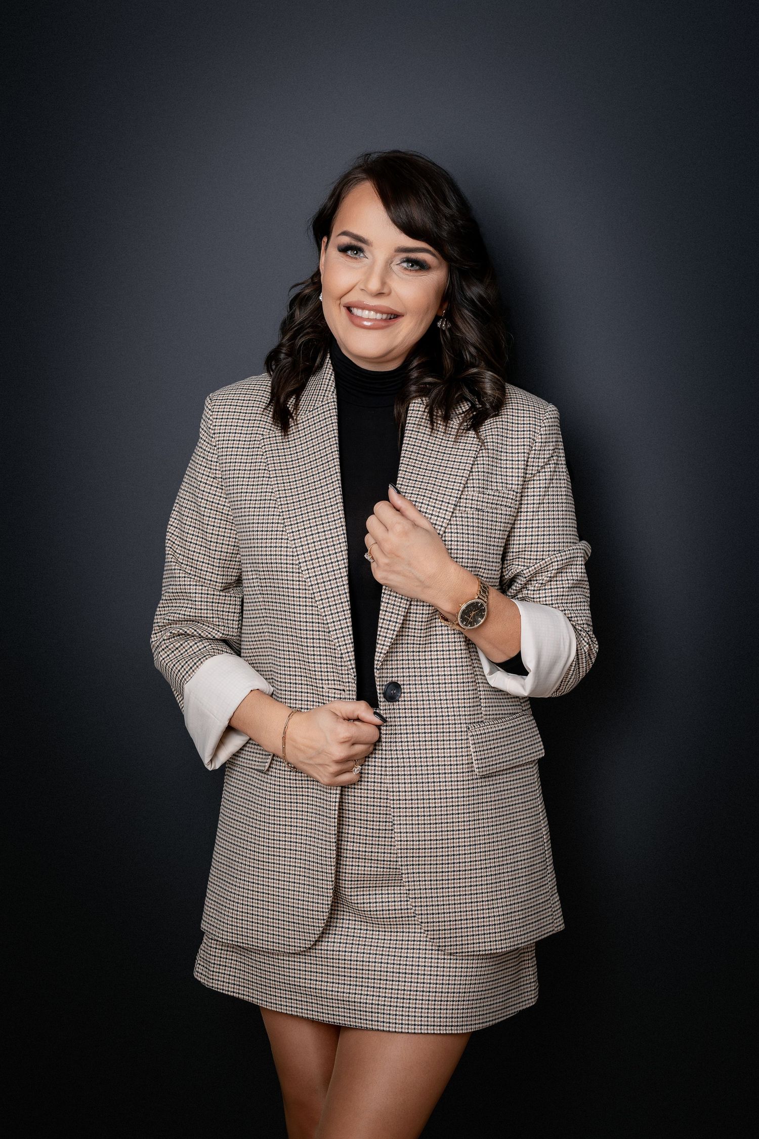 Professional studio portrait of someone in a stylish plaid blazer and skirt set against dark background.
