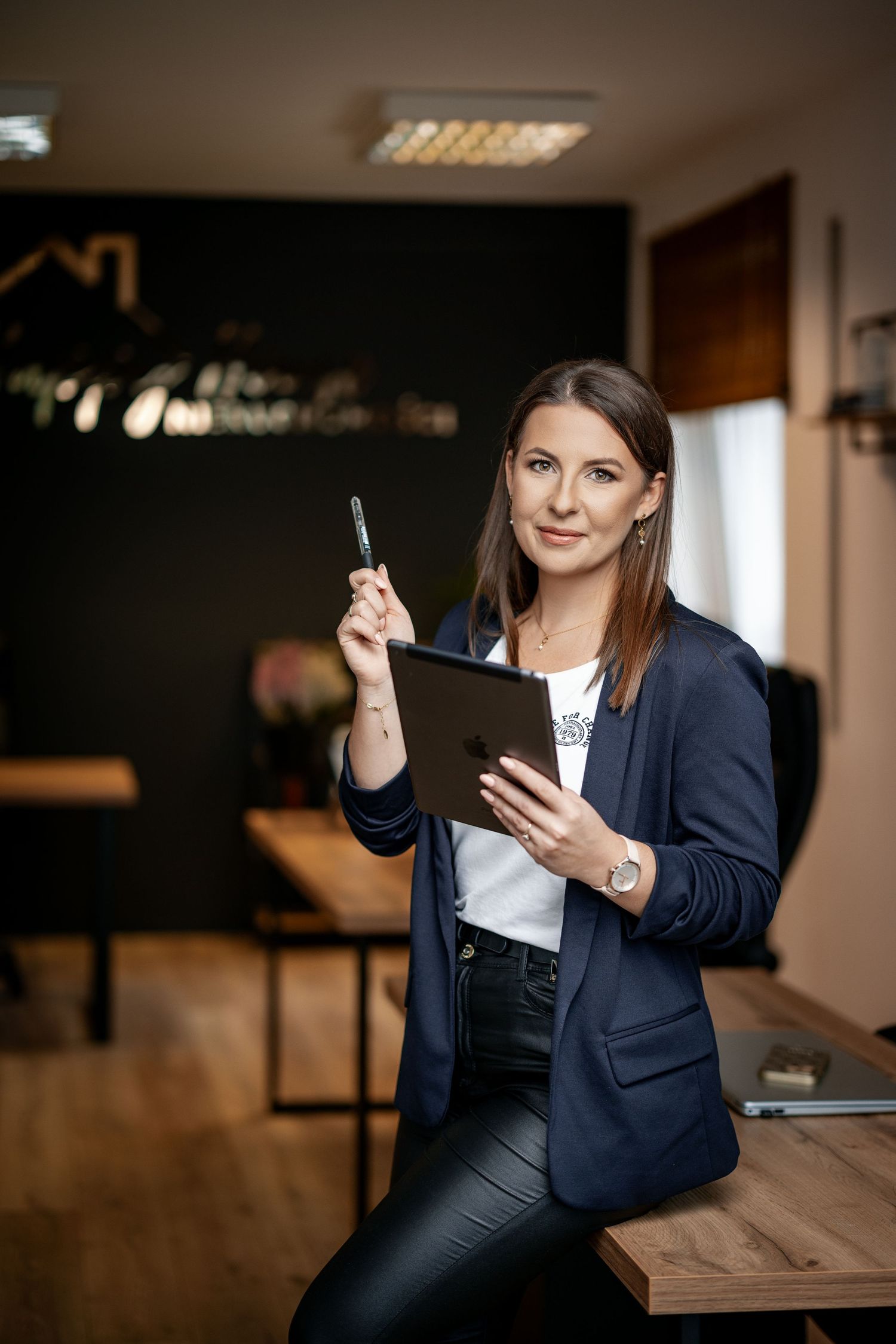 Business professional holding tablet and pen while seated on desk in modern office.