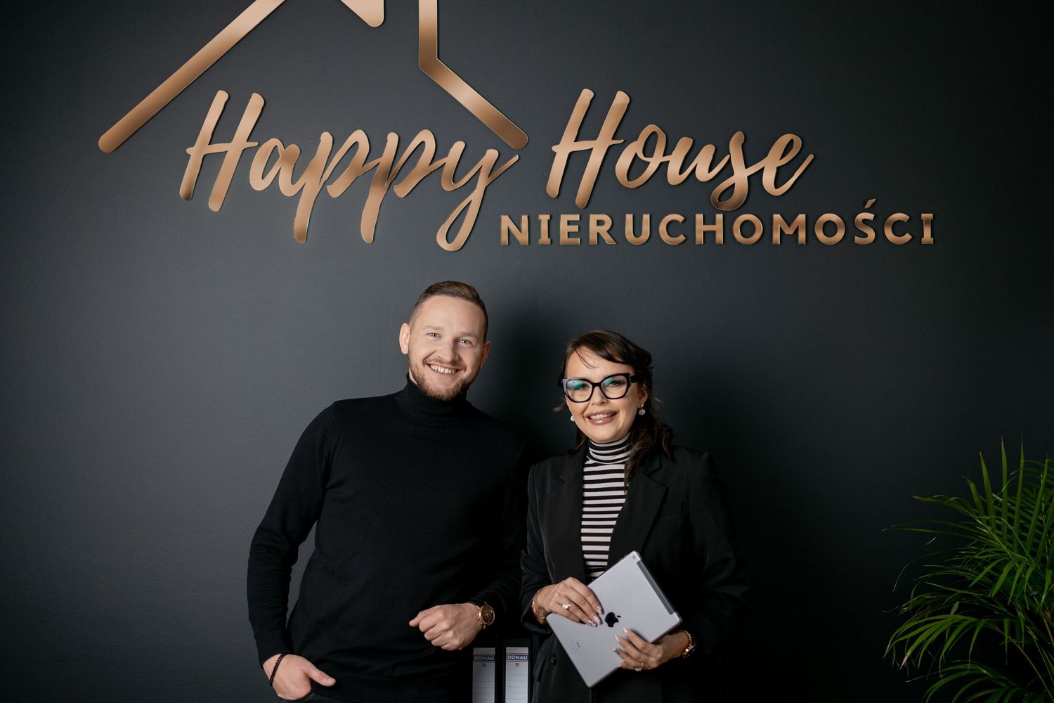 Two real estate professionals stand together in front of a dark wall with copper company branding.