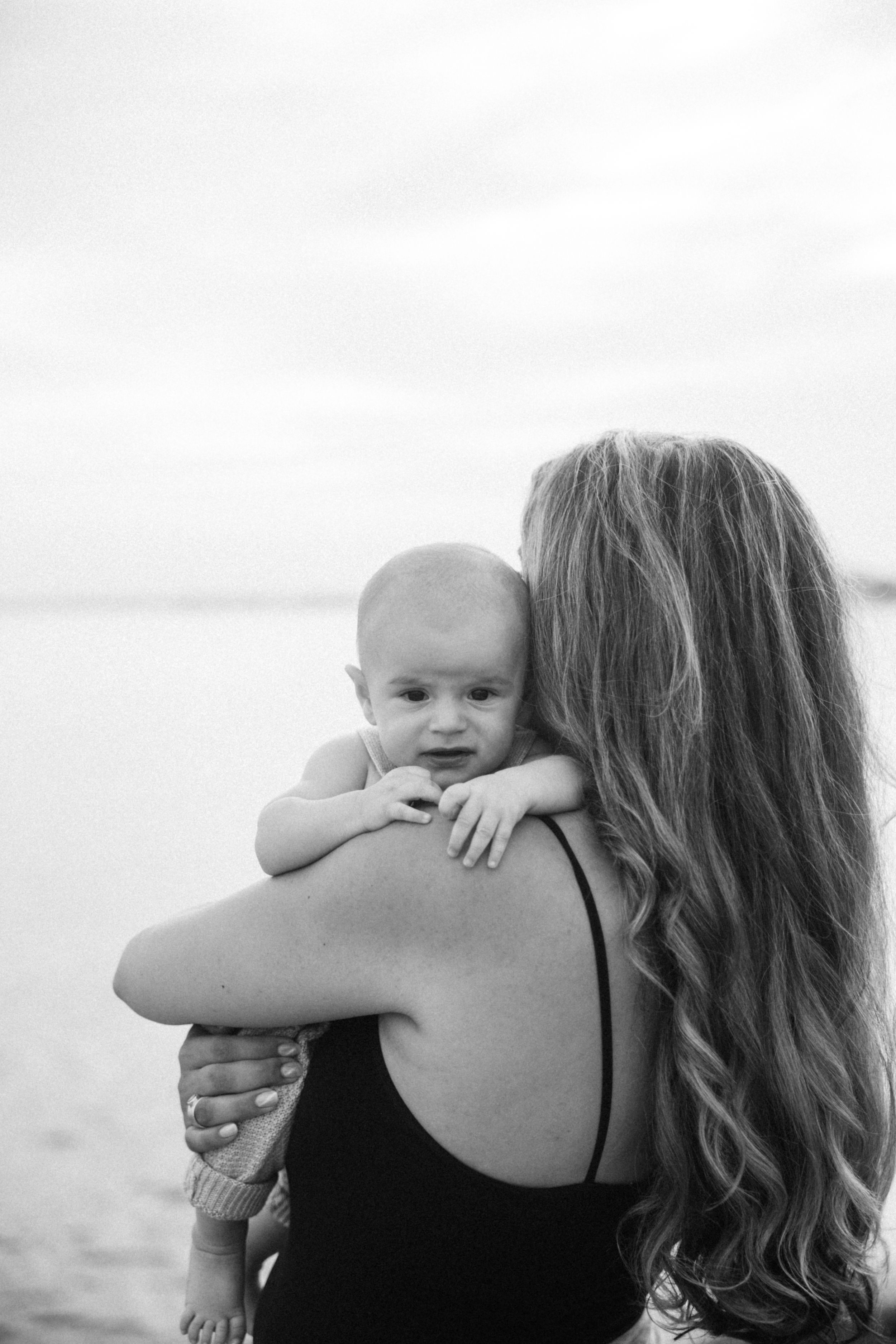 Baby smiles while being held over mother's shoulder on beach in black and white photo.