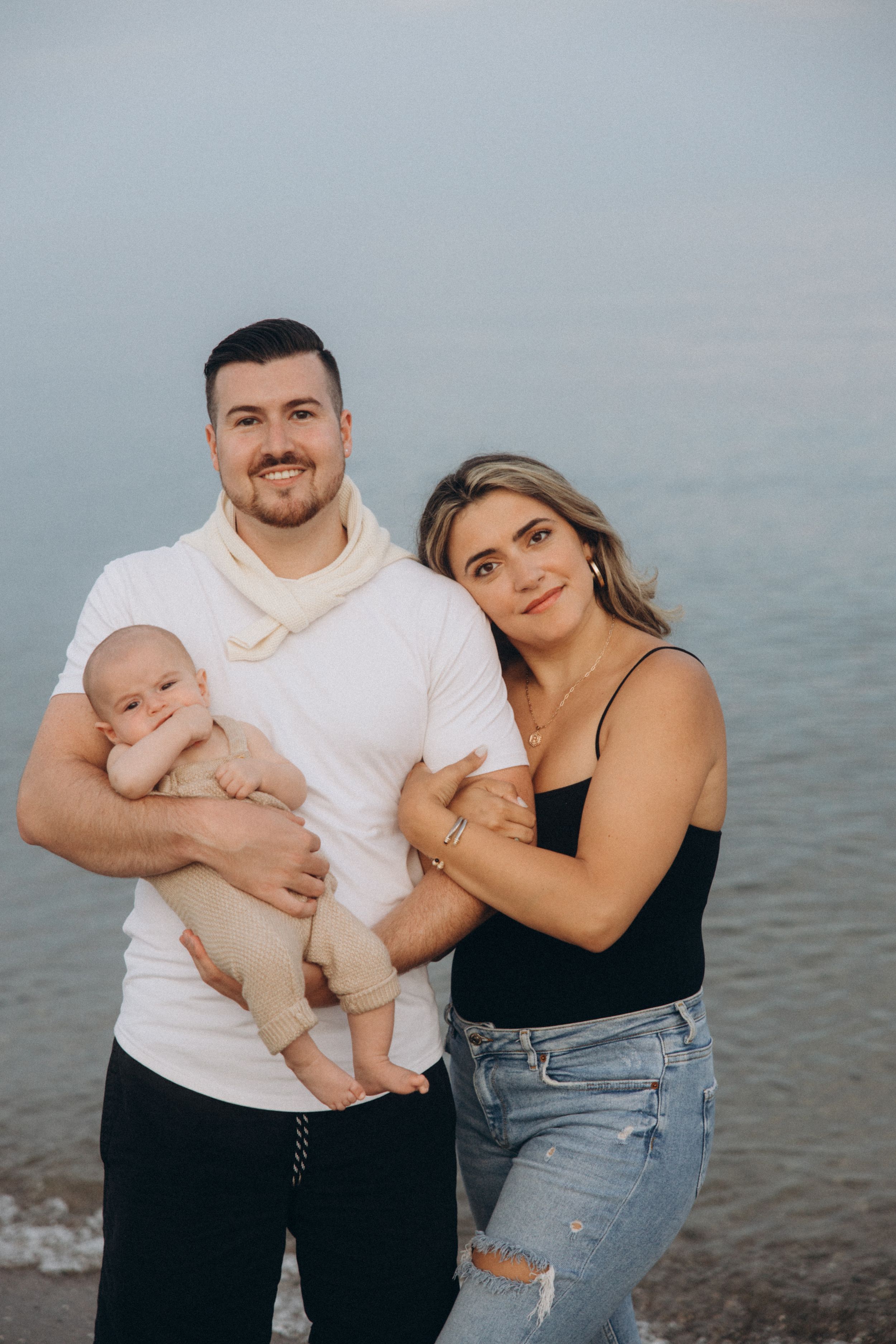 Family of three poses together on beach at sunset with calm waters in background.