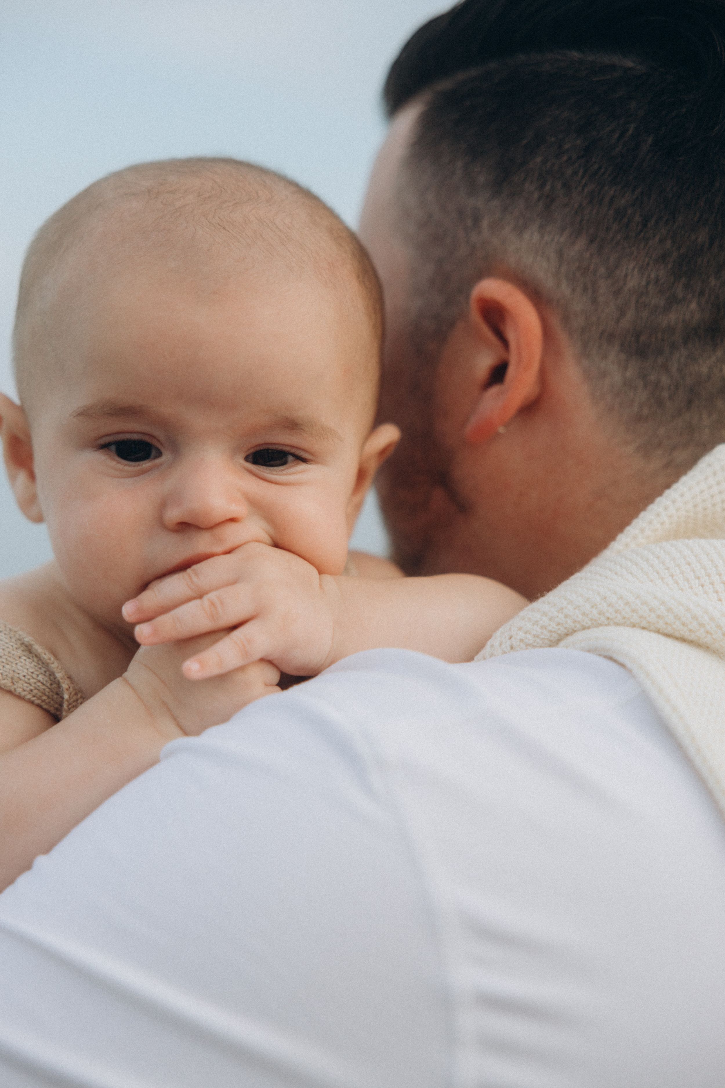 Close up shot of baby with hand near mouth being held against white shirt.