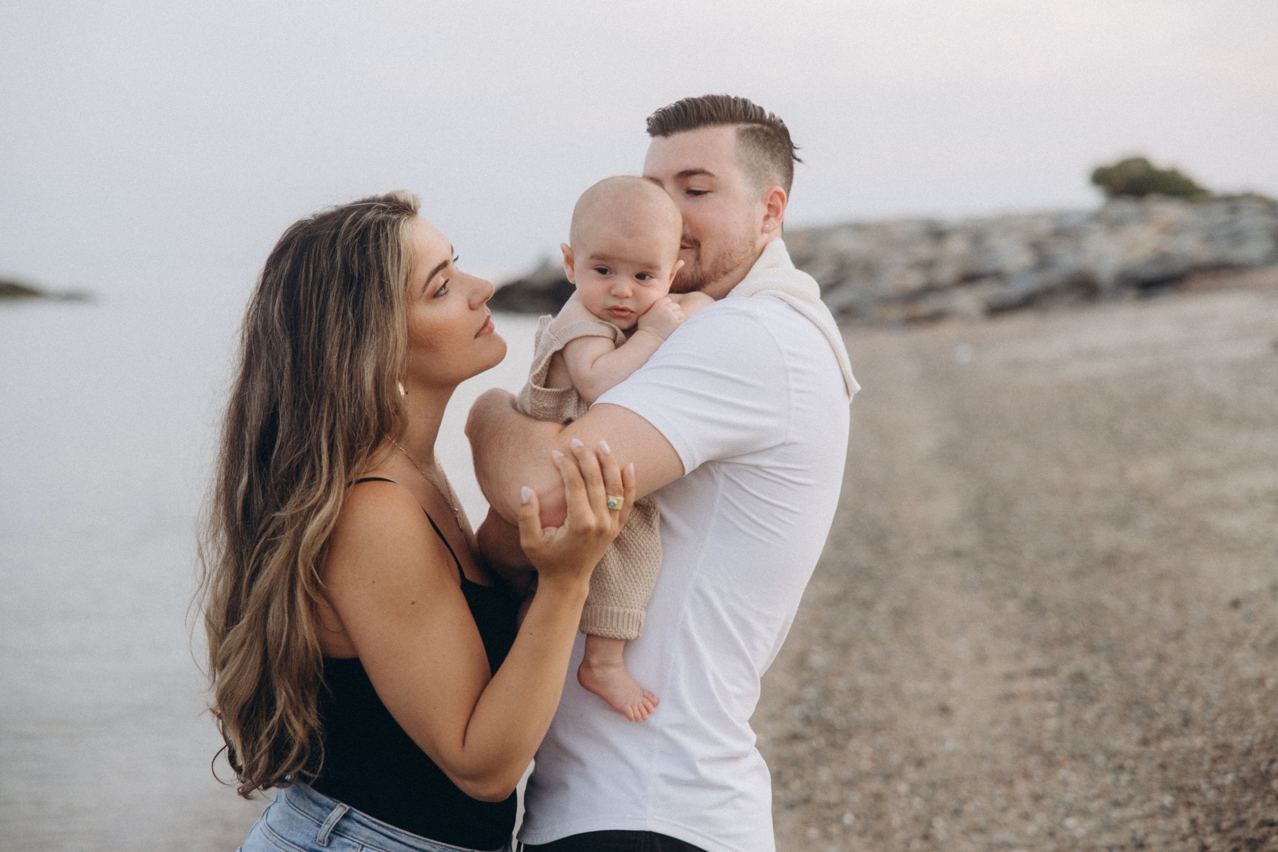 Parents sharing tender moment with their baby on beach with rocks in background.