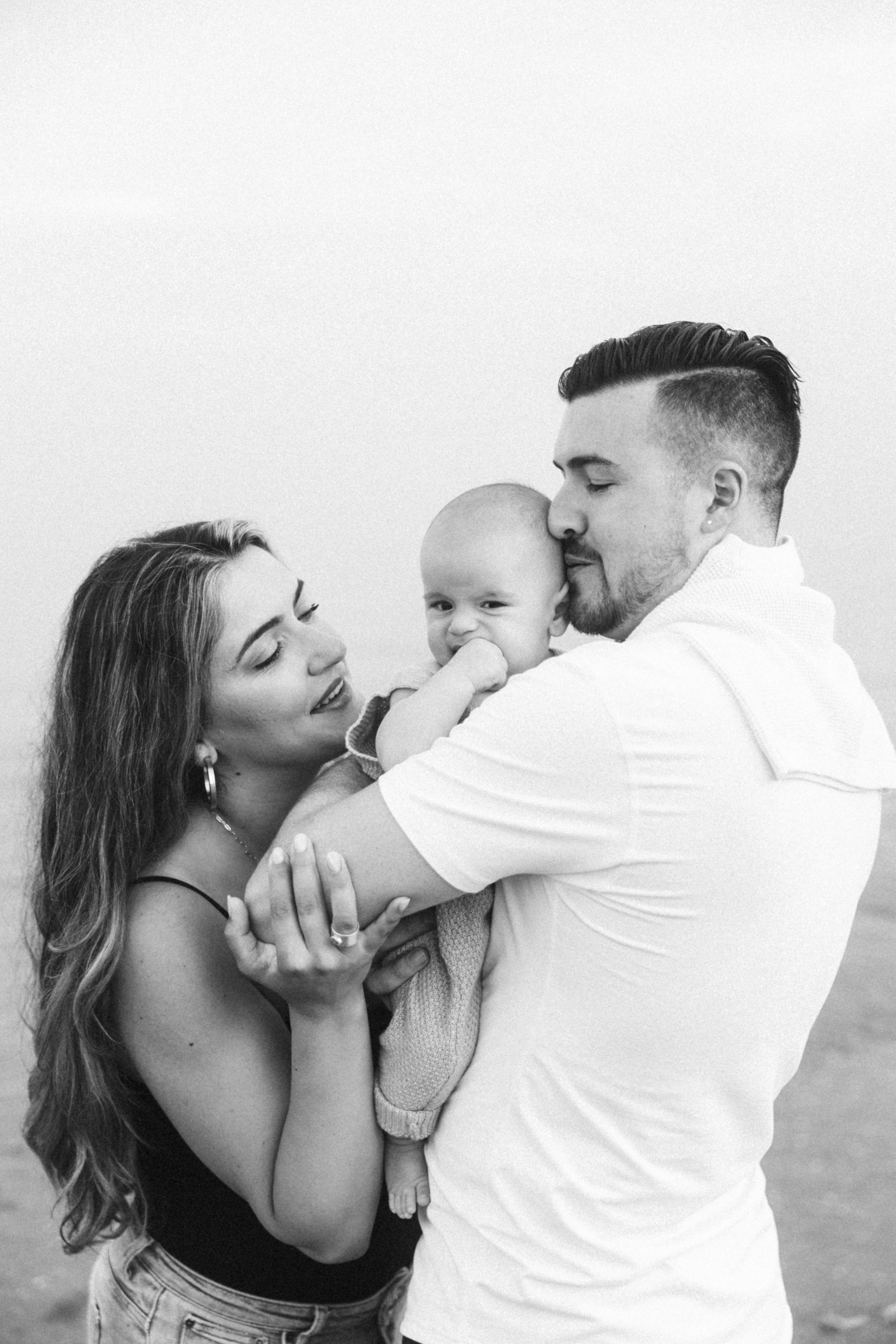 Black and white intimate family portrait showing parents embracing baby on beach.