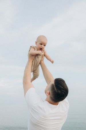 Parent lifts smiling baby in white shirt up against cloudy sky by ocean.