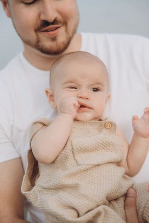 Baby wearing beige knit romper touches their nose while being held by someone in a white shirt.