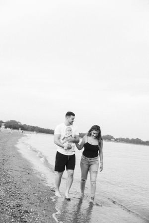 Black and white photo of a couple walking barefoot along the beach with their baby.