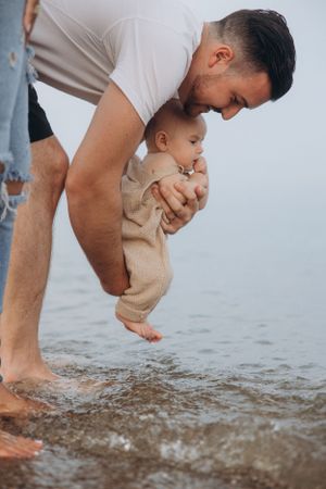 Parent holding baby in beige romper to touch the water at the beach.