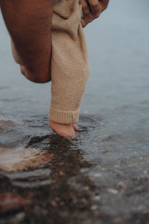 Close up of small baby feet in beige knit clothing touching water.