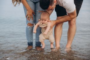 Parents helping baby touch the water at the beach while wearing beige outfit.