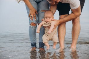 Baby in beige romper touches ocean water while supported by parents at the beach.