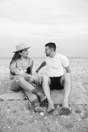 Family relaxing on beach blanket as mother in sun hat cradles infant.