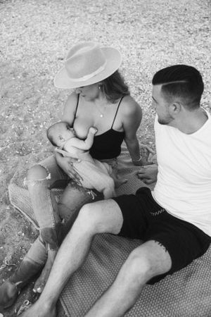 Mother in hat nursing baby while sitting with partner on beach blanket.
