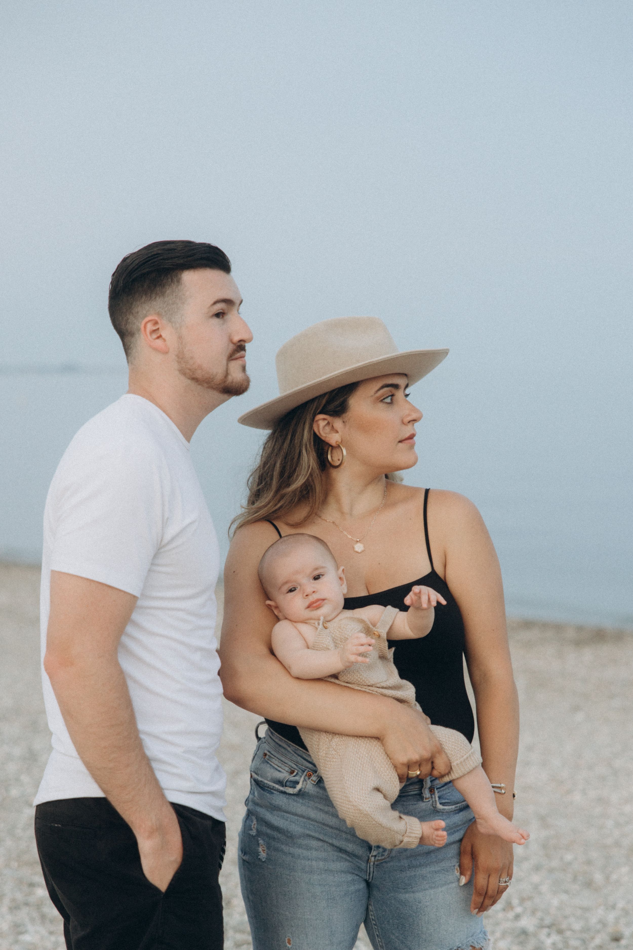Family in casual attire poses together near ocean with infant.