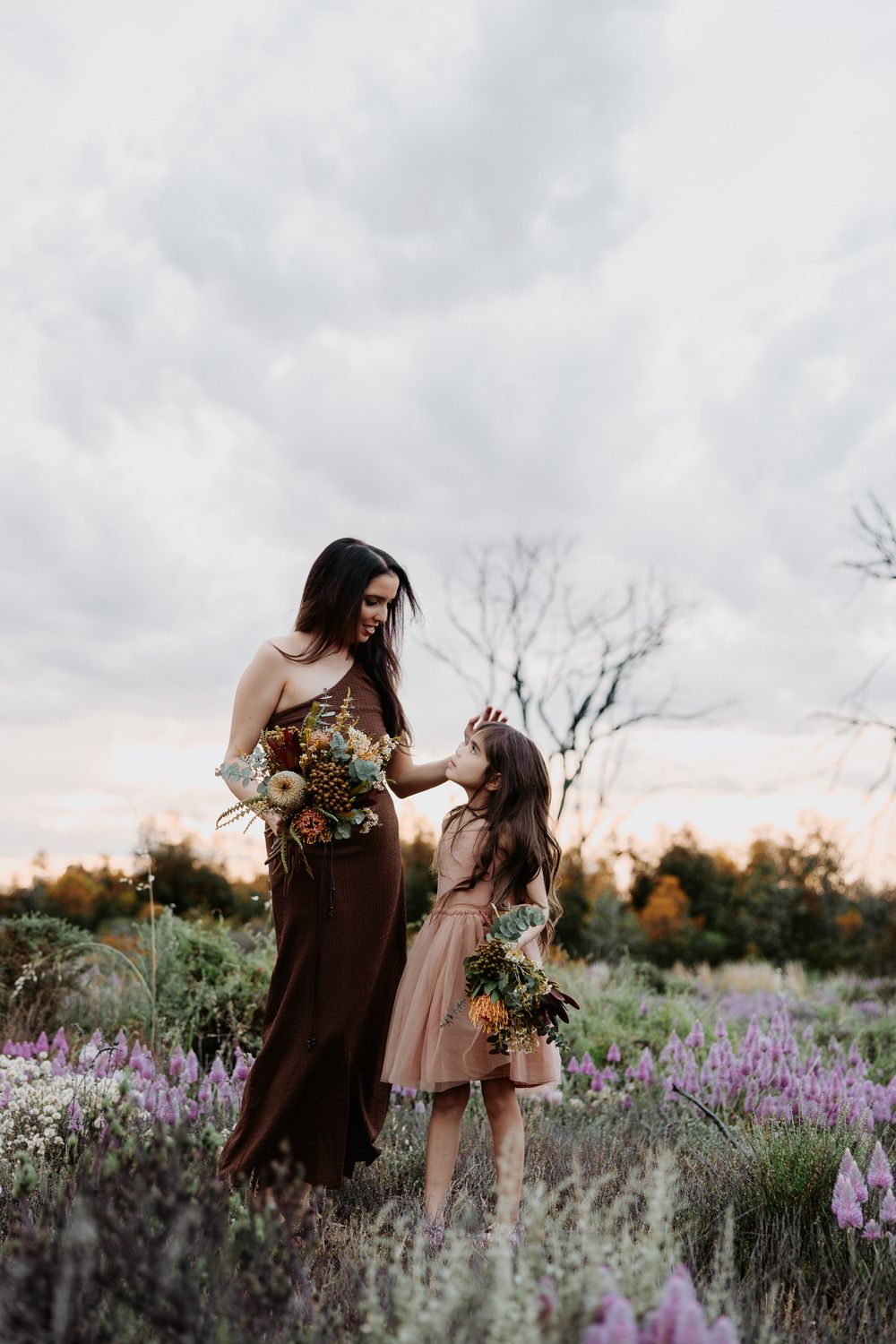 Two figures share a tender moment in a field of purple wildflowers at sunset with cloudy skies overhead.