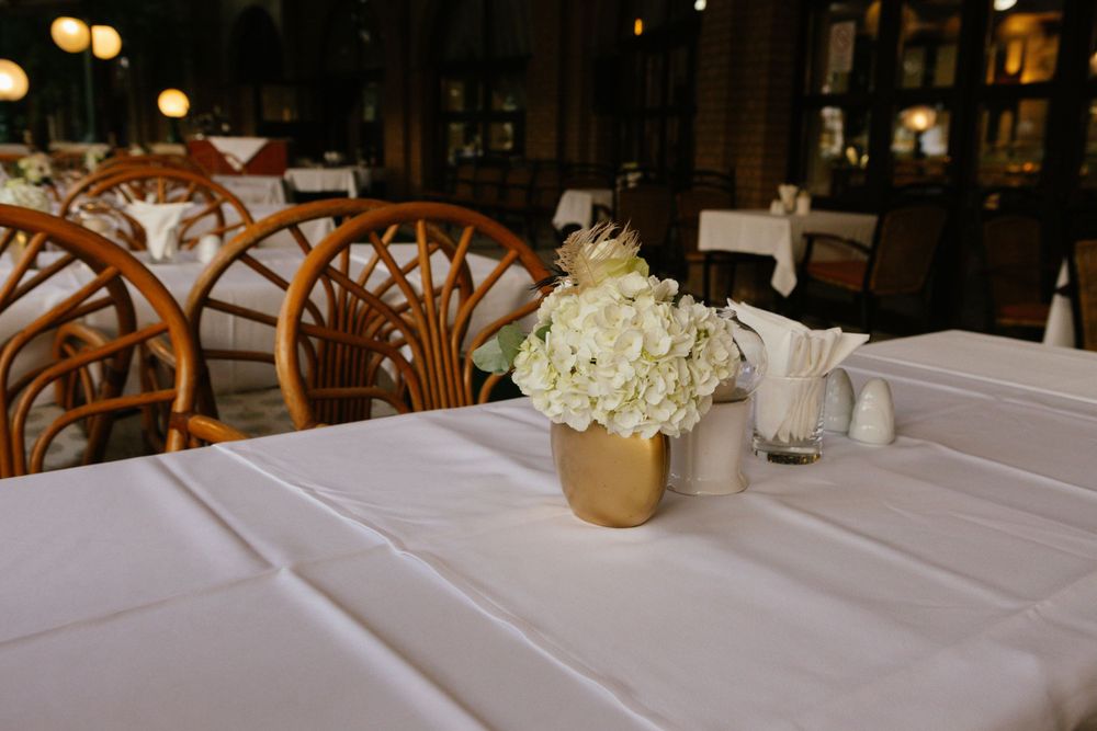 White floral centerpiece in gold vase on elegant table setting with rattan chairs in dimly lit restaurant.