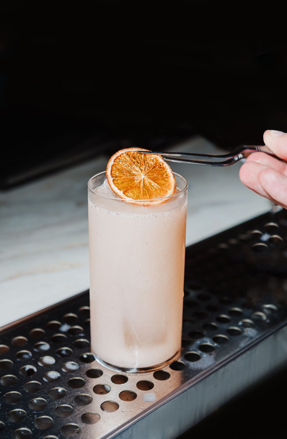 Pink cocktail garnished with dried orange slice on metal bar mat.