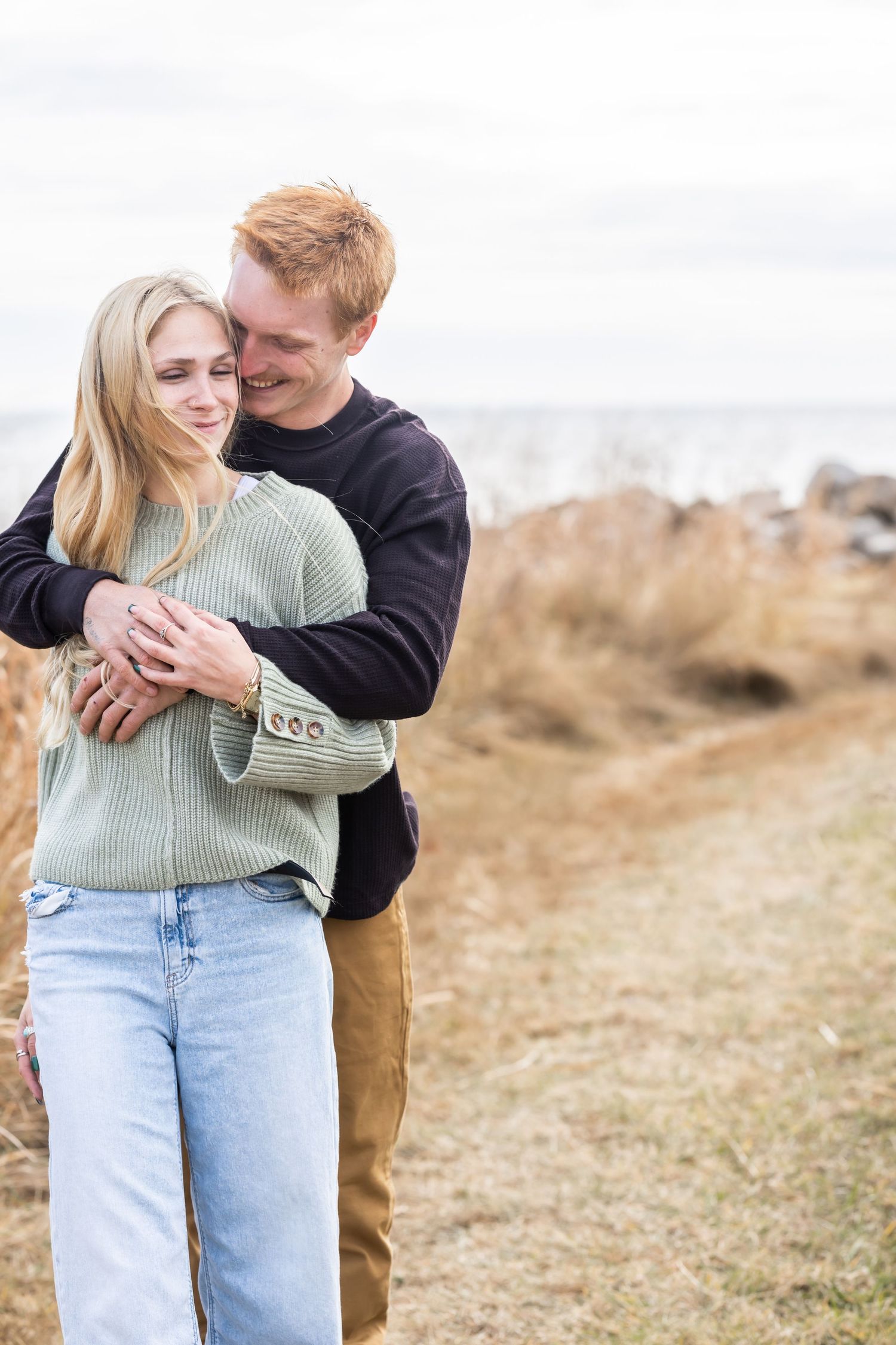 Casual couple portraits in sweaters embracing on coastal grassland during golden hour.