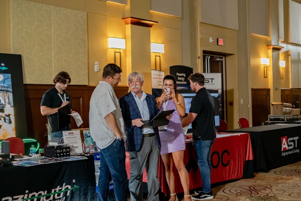 Conference room setup with vendor tables and attendees mingling in hotel setting.