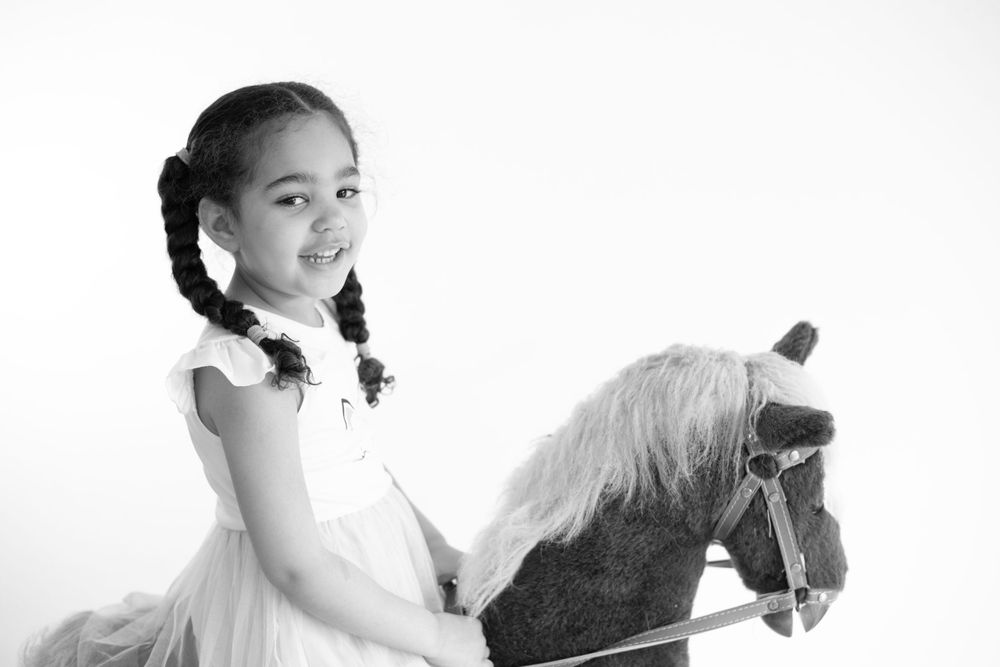 A young girl in a dress poses with a rocking horse against a white background.
