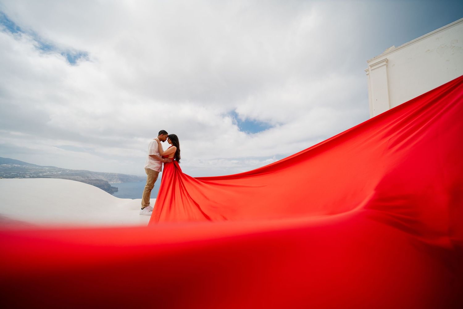 Dramatic red fabric billows against white Mediterranean architecture and cloudy blue skies.