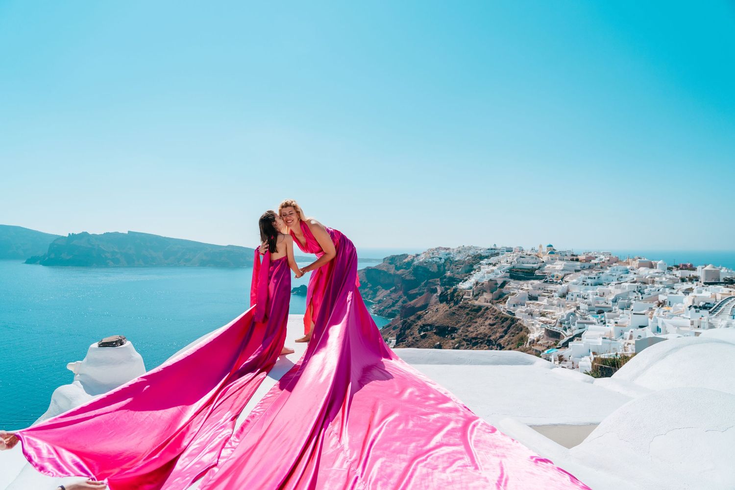 Magenta fabric billows dramatically on white rooftop with Santorini's coastline in background.