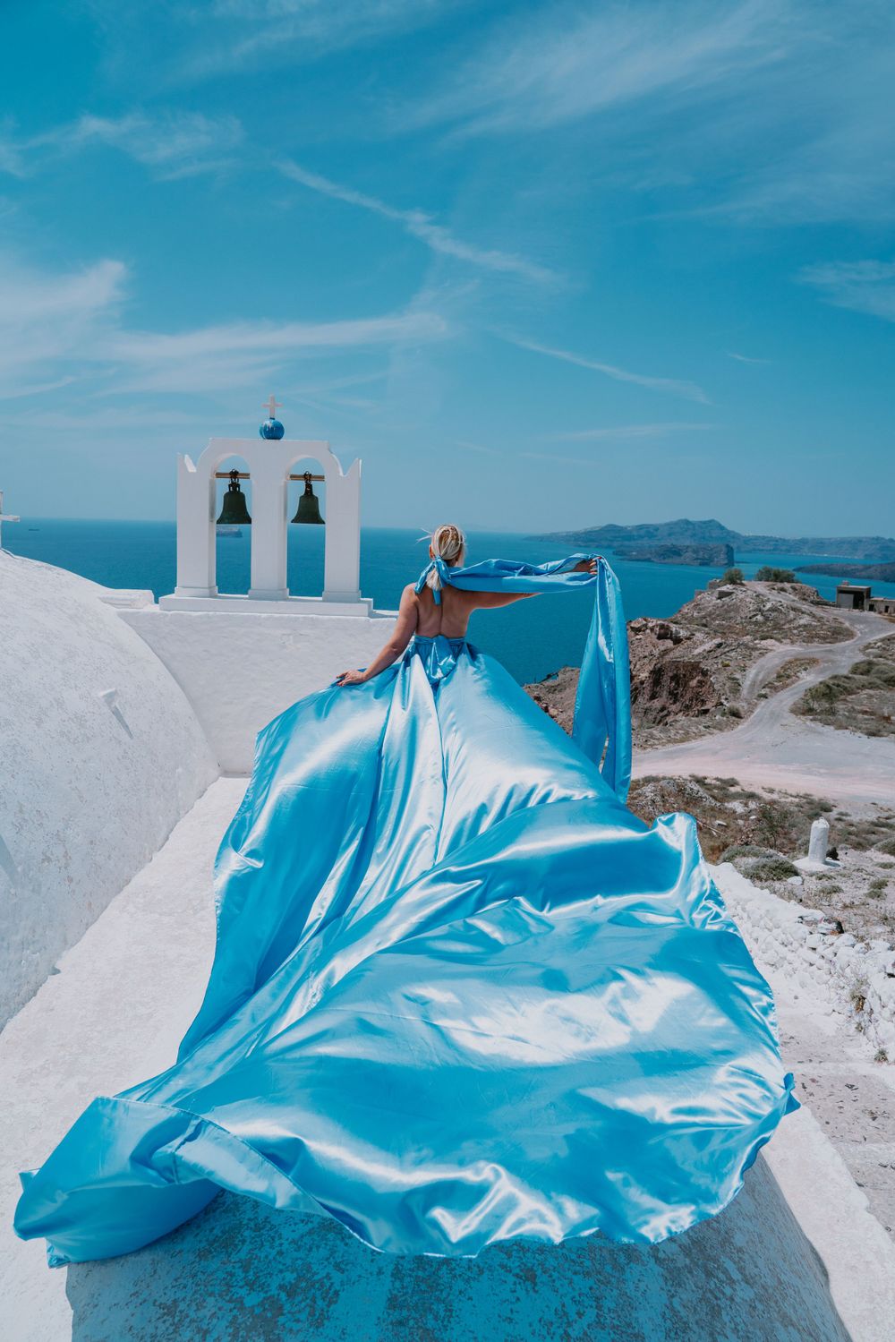 A flowing blue dress billows in the wind against white Santorini architecture with traditional bell towers and ocean views.