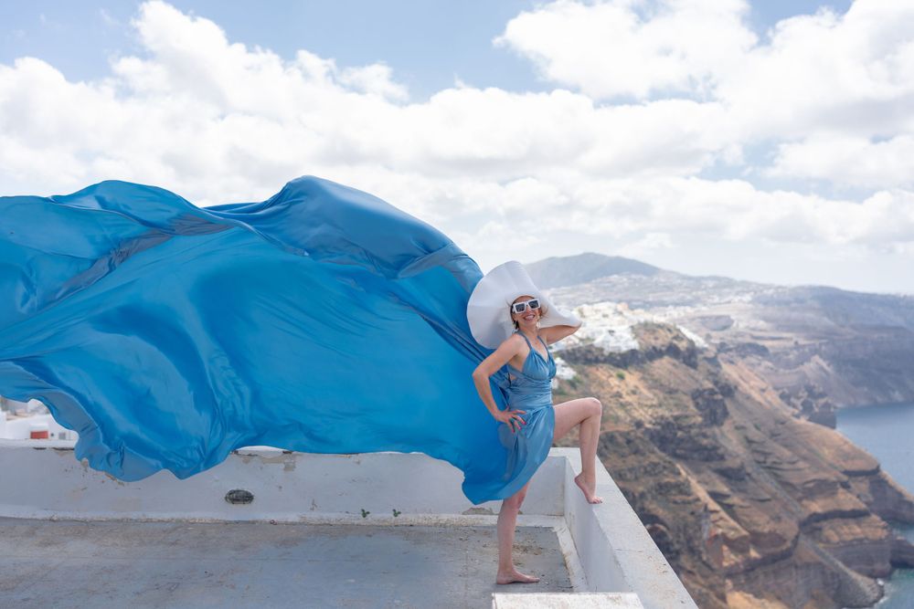 Flowing blue dress fabric billows dramatically against Santorini cliff landscape.