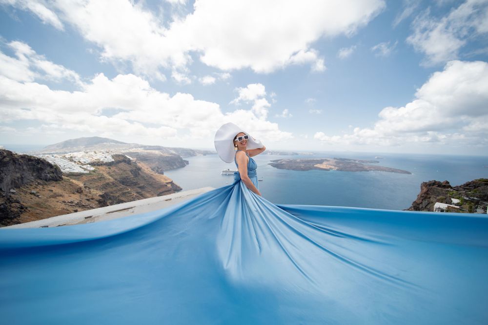Flowing light blue dress billowing dramatically against Santorini's coastal landscape and blue sky.