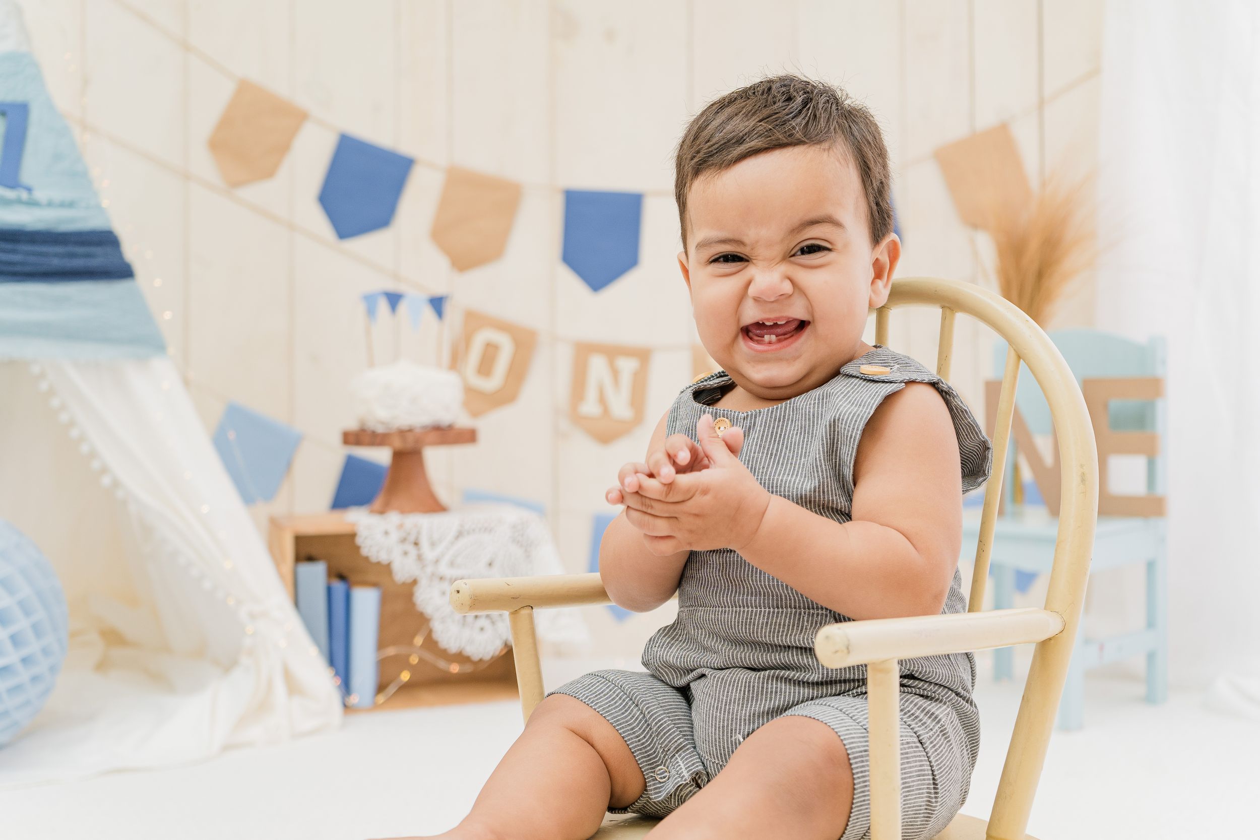 Smiling toddler sitting in wooden chair with party decorations in background.