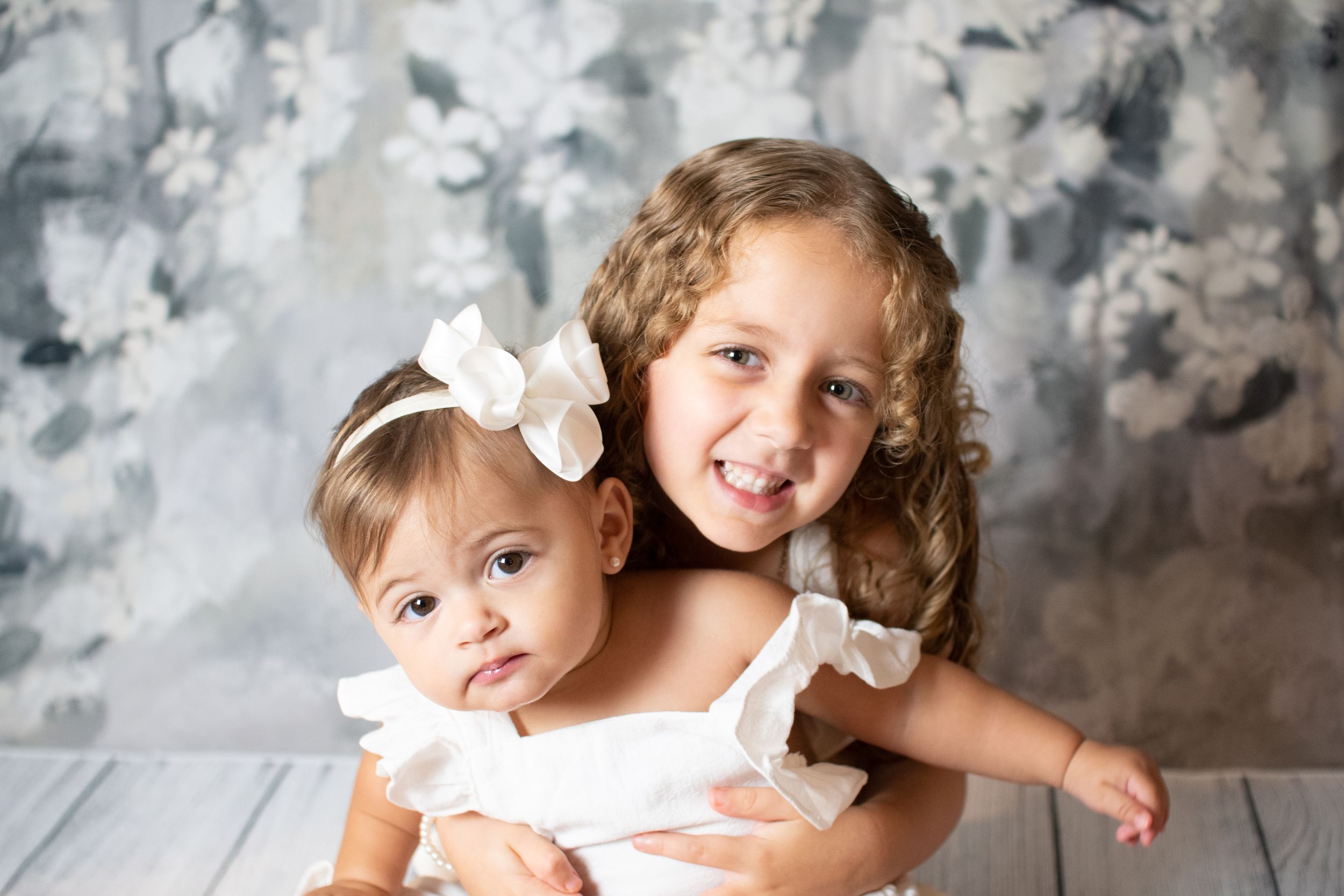 Two children in white dresses with hair bows pose against gray textured background.