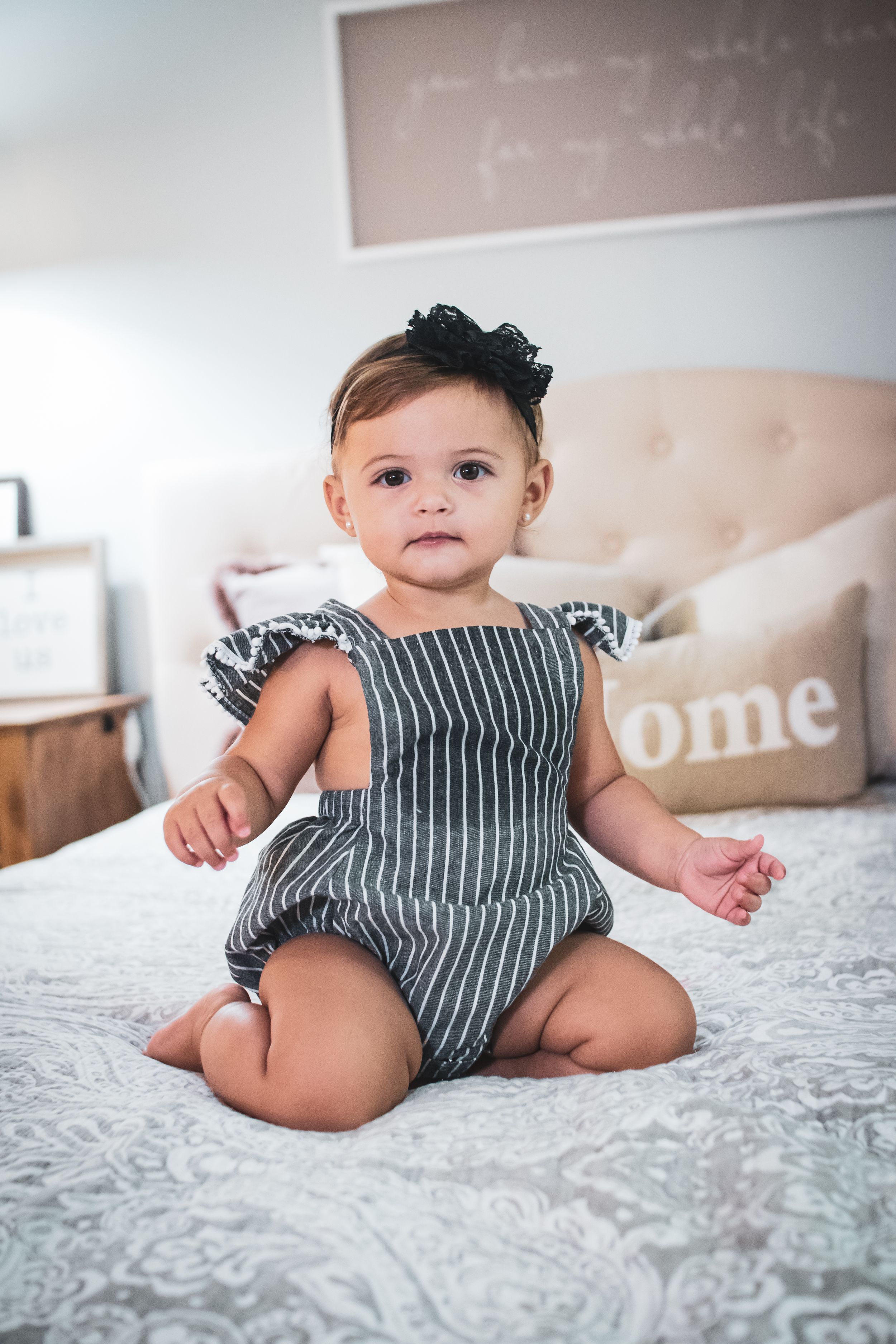 Baby wearing striped romper sits on white bedding with decorative pillow in background.