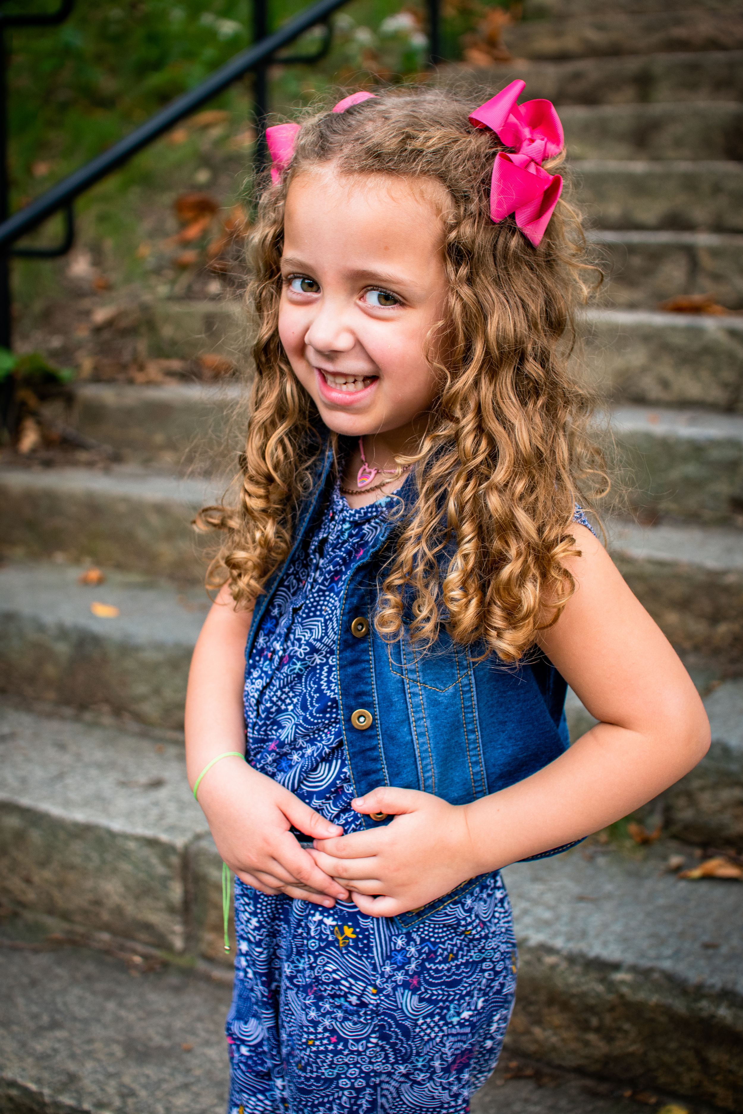 Young girl in blue dress with pink bow smiling on outdoor stone steps.