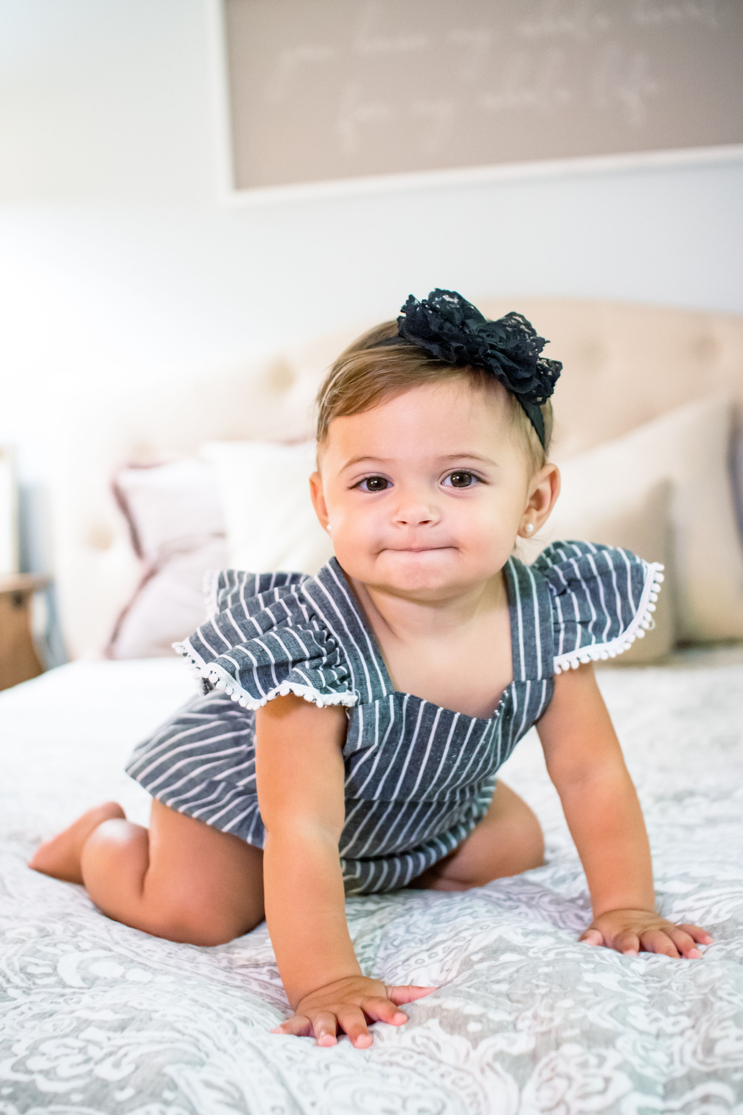 Baby in striped romper and bow crawling on white bedding.