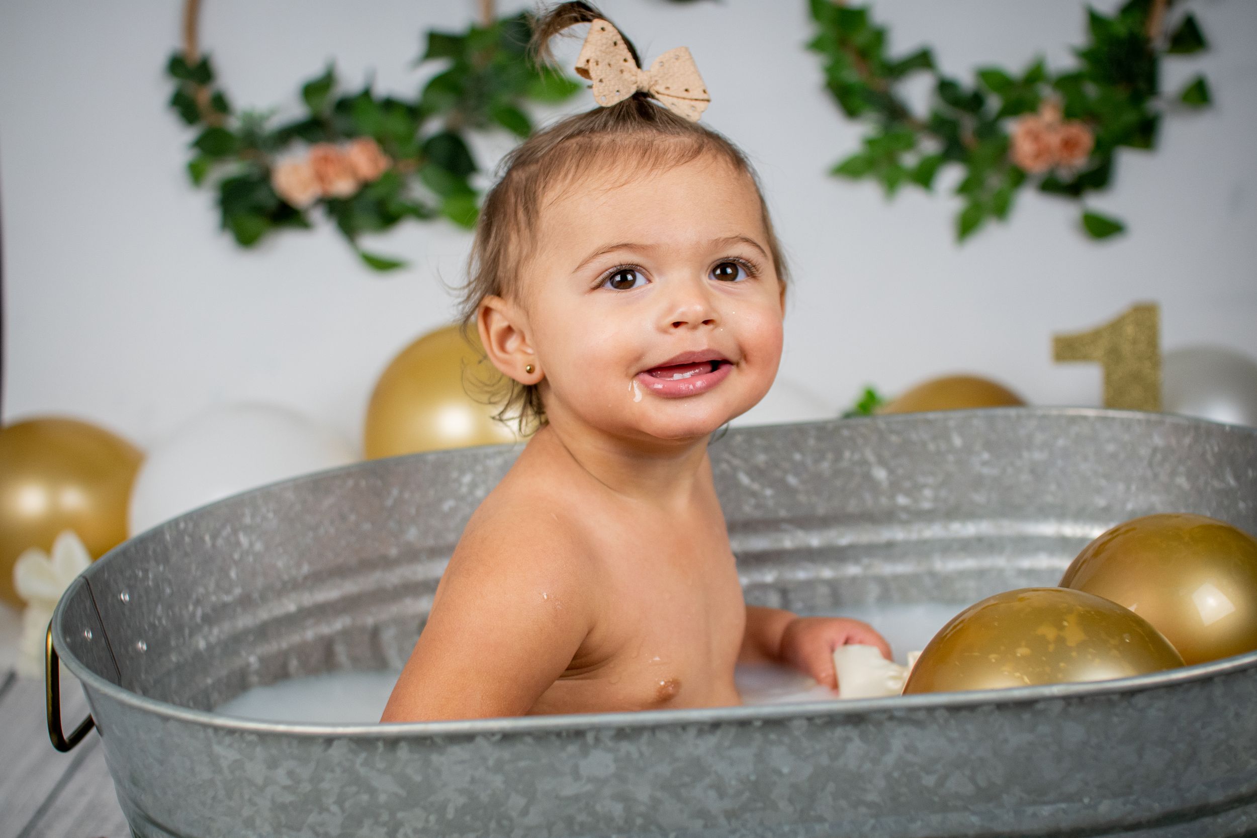 Happy baby taking bath in metal tub with gold balloons and greenery backdrop.